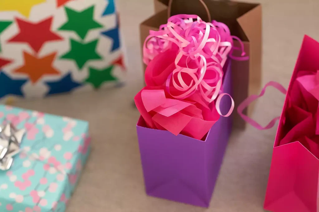 Brightly colored gift bags filled with pink tissue paper and curly ribbons, surrounded by wrapped presents in festive designs